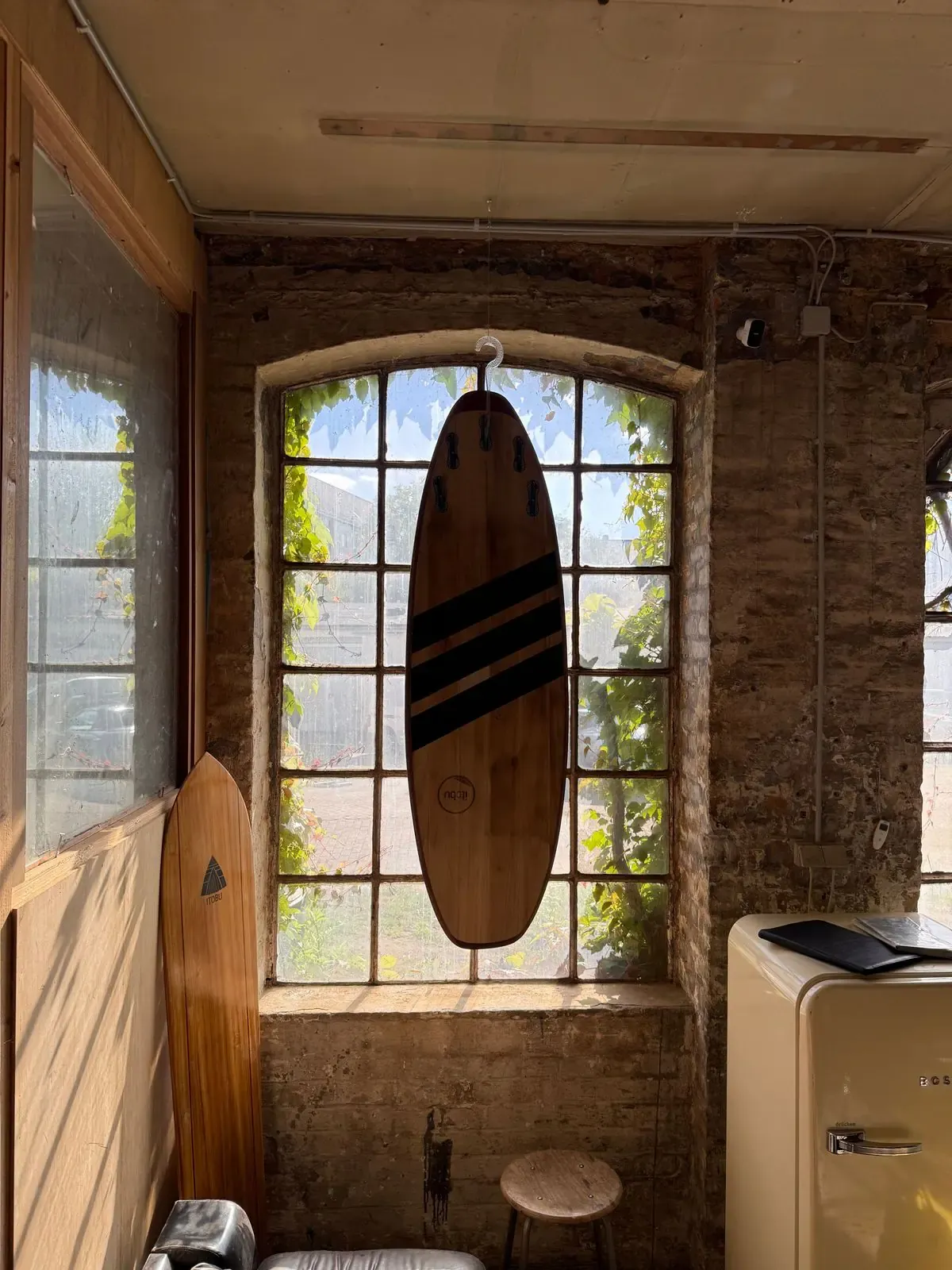 Surfbrett aus Holz mit drei dunklen Streifen und Finnen aus Holz, aufgehängt vor einem Rundbogenfenster